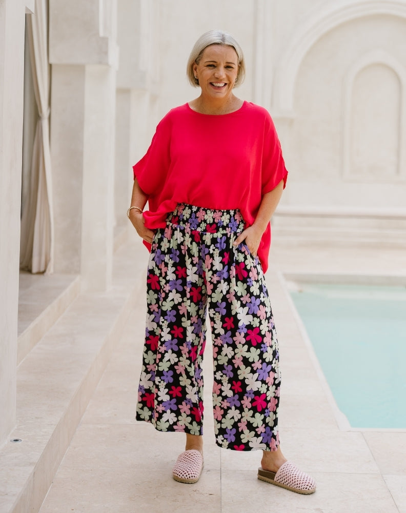 A woman stands outdoors next to a light-colored pool, wearing a bright red short-sleeve top and high-waisted, wide-leg floral pants with pink, purple, and white flowers on a dark background. She is wearing pink-and-white checkered slip-on shoes.