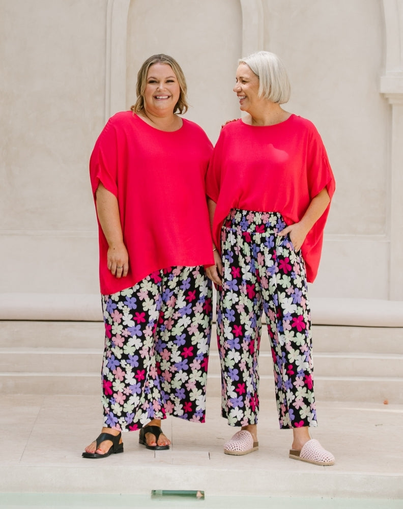 Two women standing side-by-side wearing bright pinkish-red short-sleeve t-shirts and wide-leg floral pants with a black, white, pink, and purple pattern.