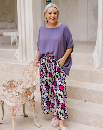 A smiling woman stands on stone steps wearing a loose lavender top and high-waisted black wide-leg pants with a colorful floral print. She is posed next to a distressed white chair.