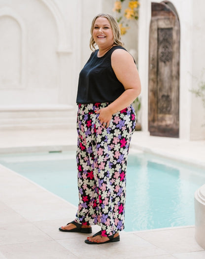 A woman modeling a sleeveless black top and high-waisted, wide-leg floral pants with a black base and pink, purple, and white flowers, standing next to a swimming pool.