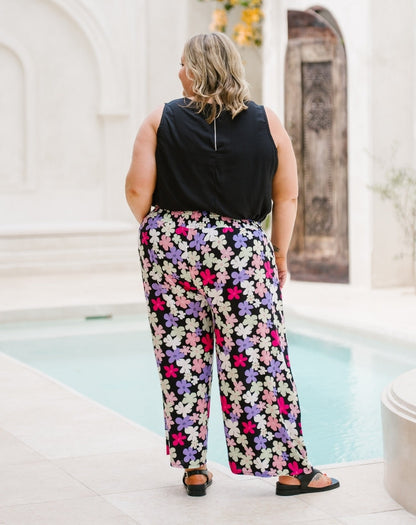 A woman viewed from behind wearing a black sleeveless top and wide-leg pants with a colorful floral print in pink, purple, white, and green, standing by a swimming pool.