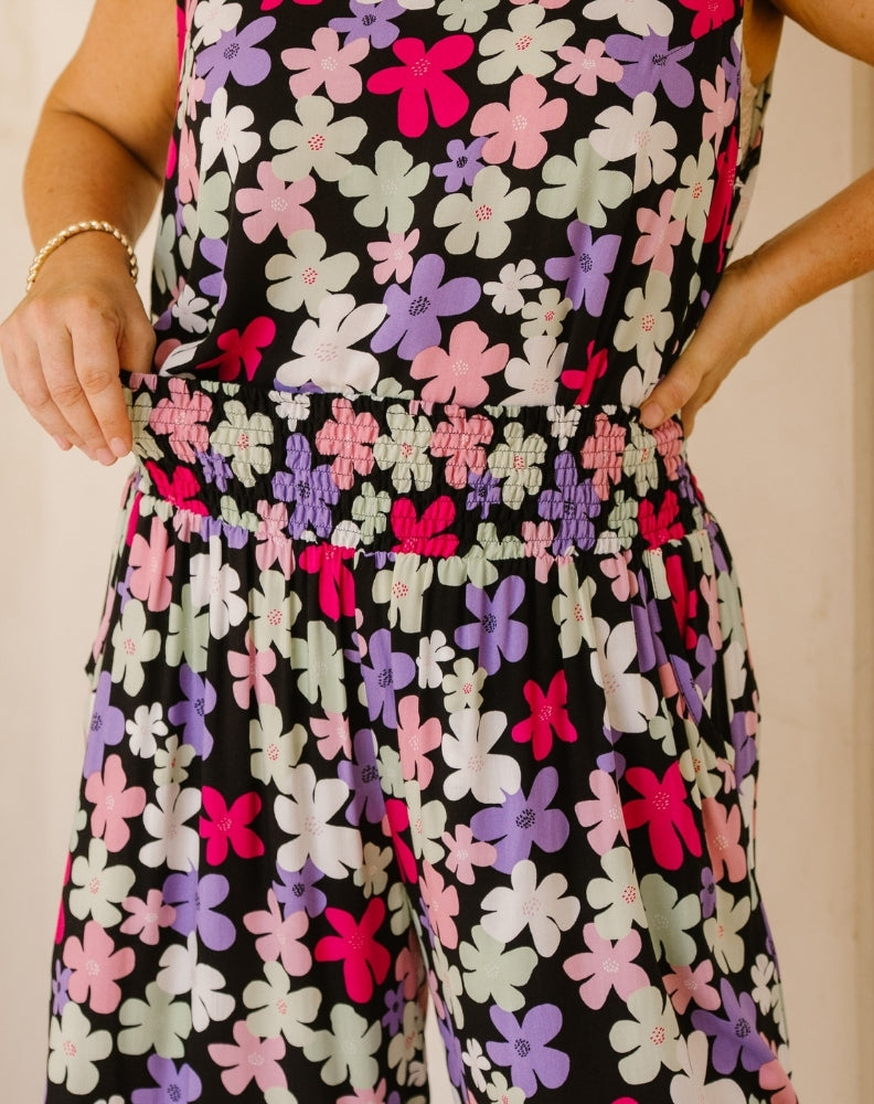 A close-up of a sleeveless jumpsuit with a black background and a colorful floral pattern of pink, purple, and light green flowers. The jumpsuit features a smocked elastic waistband and is modeled by a person whose hands and arms are visible.