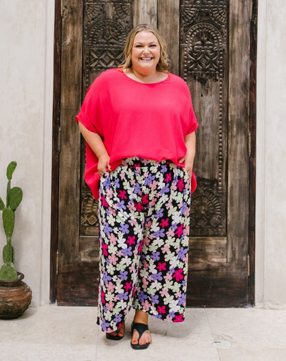 A woman modeling a bright pink short-sleeve top and black wide-leg pants with a colorful floral print, standing in front of a rustic dark wooden door.