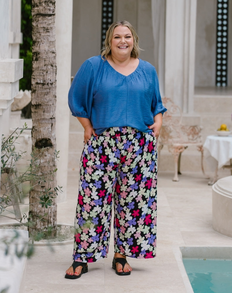 A woman stands outdoors in a courtyard wearing a loose blue blouse and wide-leg floral pants with black sandals.