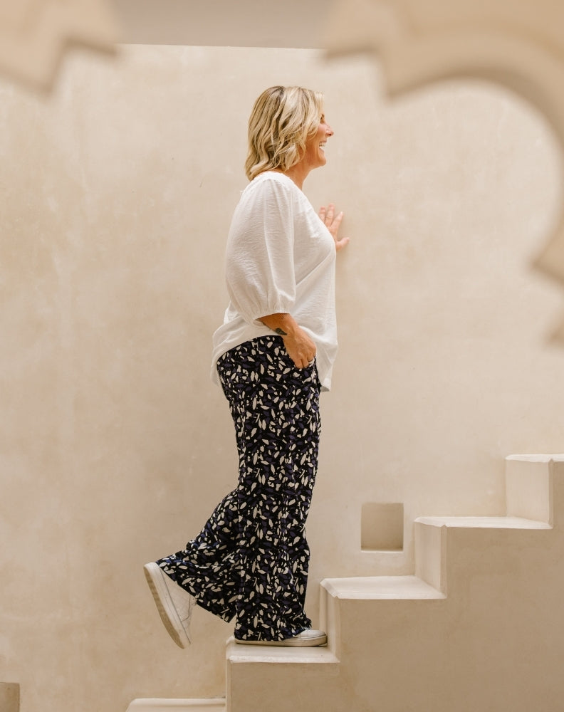 A woman wearing a white textured blouse and black wide-leg pants with a white leaf pattern, walking down white steps against a warm beige stucco wall.