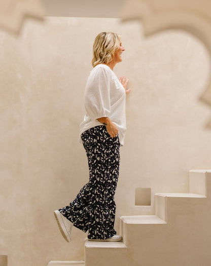 A woman wearing a white textured blouse and black wide-leg pants with a white leaf pattern, walking down white steps against a warm beige stucco wall.