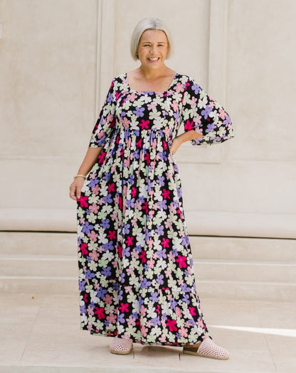A woman wearing a long-sleeved, floor-length floral maxi dress with a black base and pink, purple, and white flowers, standing on stone steps.