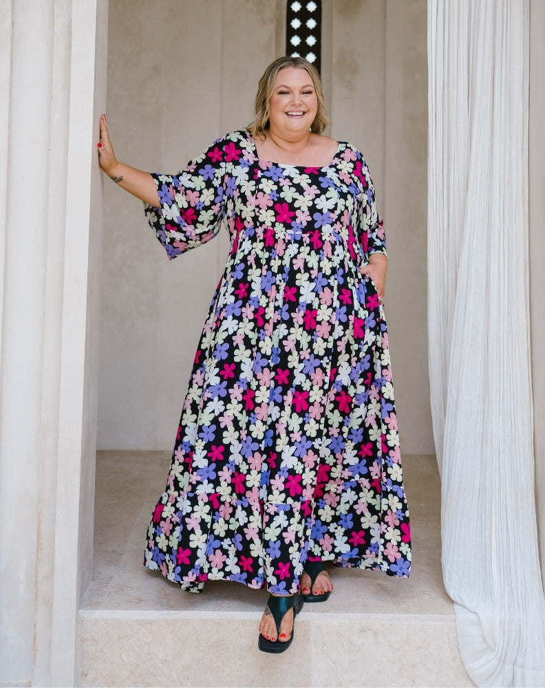 A woman modeling a long-sleeve, floor-length floral maxi dress with a black background and pink, purple, and white flowers, standing in a doorway with white drapes.