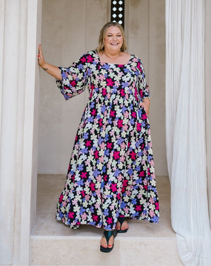 A woman modeling a long-sleeve, floor-length floral maxi dress with a black background and pink, purple, and white flowers, standing in a doorway with white drapes.