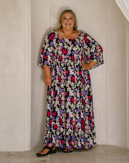 A woman modeling a long-sleeved, floor-length maxi dress with a dense multicolor floral print on a black background, standing against a cream-colored paneled wall.