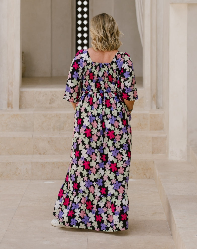 A woman viewed from behind wearing a black maxi dress with a colorful floral pattern of pink, purple, and white flowers, standing on stone steps in a classical building.