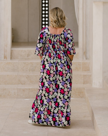A woman viewed from behind wearing a black maxi dress with a colorful floral pattern of pink, purple, and white flowers, standing on stone steps in a classical building.