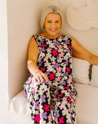 A smiling woman with short blonde hair sits on a white cushioned bench wearing a sleeveless black jumpsuit covered in a colorful floral pattern of pink, purple, white, and light green flowers.