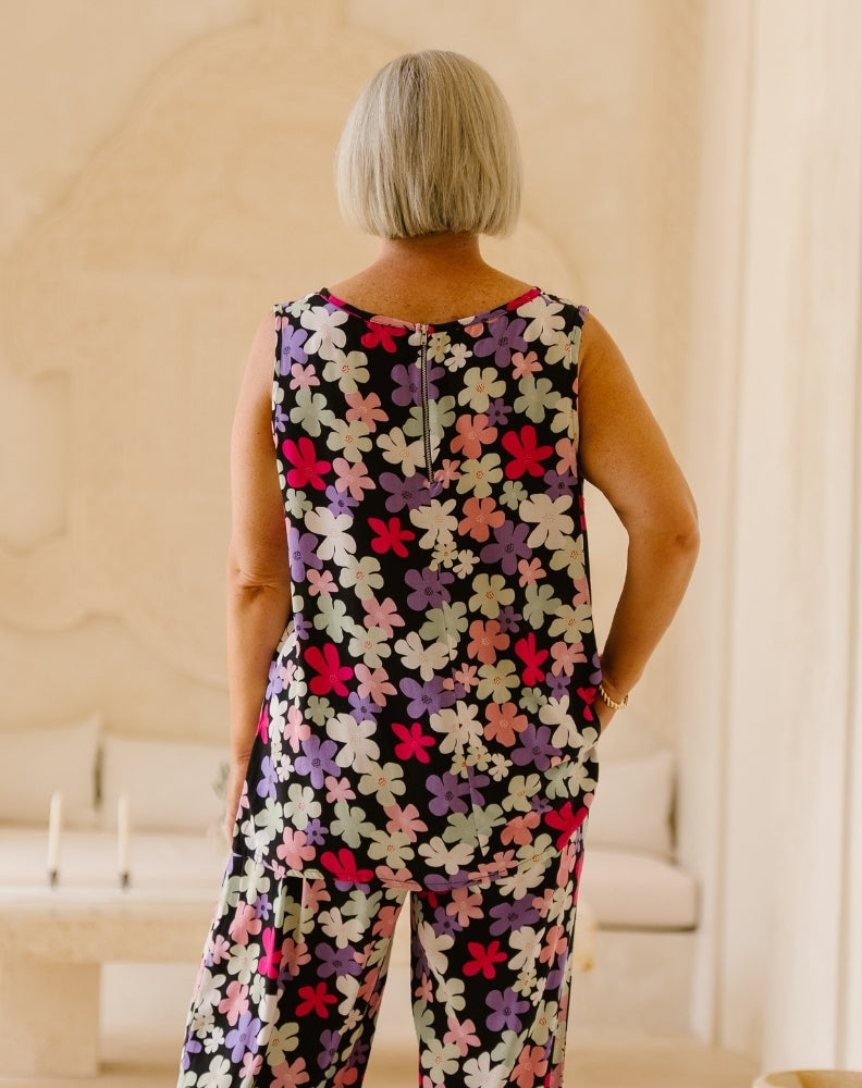 A woman with a short blonde bob is shown from behind wearing a sleeveless jumpsuit with a colorful floral pattern on a black background, standing in a brightly lit indoor setting.