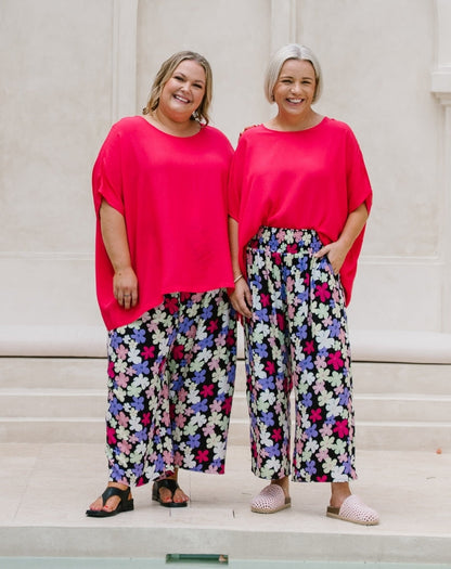 Two women standing side-by-side wearing matching outfits consisting of a bright pink short-sleeve top and wide-leg floral pants. The pants feature a multicolor pattern of pink, purple, blue, and white flowers on a dark background.