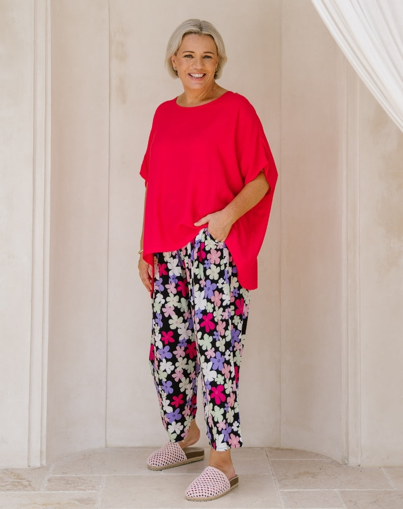 A smiling older woman stands indoors wearing a bright red oversized short-sleeve top and loose black pants with a colorful floral print in pink, purple, white, and blue. She is wearing pink-and-white checkered slip-on shoes.