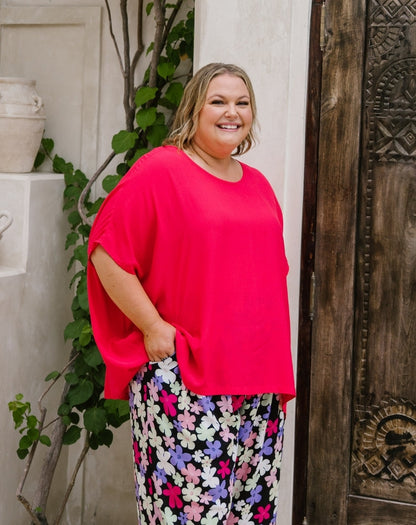 A woman wearing a bright pink short-sleeve top and a black pants with a colorful floral pattern, standing in front of a rustic wooden door and a white wall with green vines.