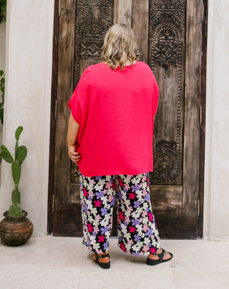 A woman viewed from behind wearing a bright pink oversized short-sleeve top and wide-leg floral print pants with black sandals, standing in front of a dark carved wooden door.