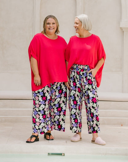 Two women standing side by side wearing matching bright pink short-sleeve tops and wide-leg floral pants with black, white, purple, and pink flowers on a light beige background.