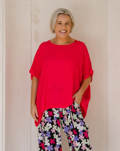 A smiling woman with short blonde hair wearing a bright red oversized short-sleeve top and black pants with a colorful floral print, standing against a light beige wall.