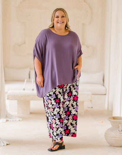 A woman modeling a two-piece lounge set featuring a loose, oversized purple short-sleeve top and matching wide-leg pants with a colorful floral print, standing in a bright, neutral-toned indoor setting.