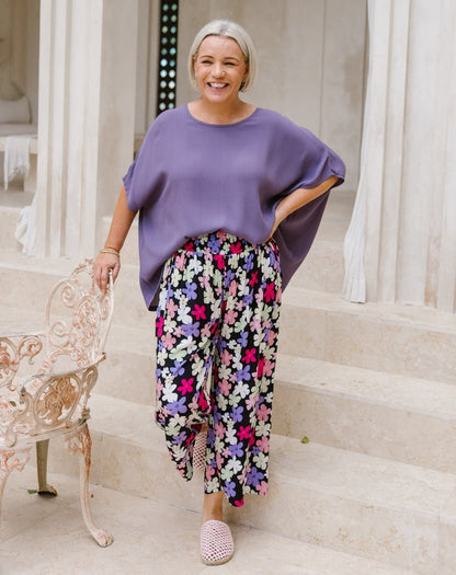 A smiling woman stands on stone steps wearing a loose lavender top and colorful floral wide-leg pants, with pink knit slip-on shoes. She is posed beside a decorative white chair in front of a light stone building.