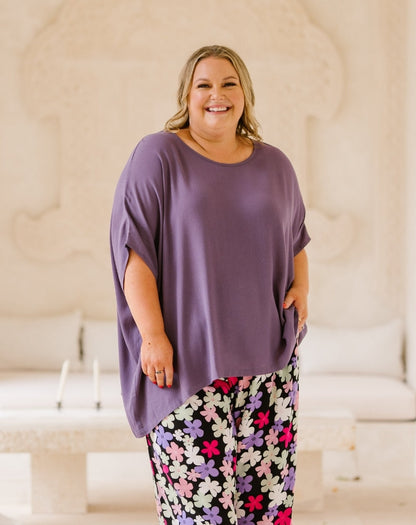 A smiling woman wearing a loose, short-sleeve mauve-purple top and black floral pants with pink, purple, and white flowers, standing in a brightly lit cream-colored room with a stone bench in the background.