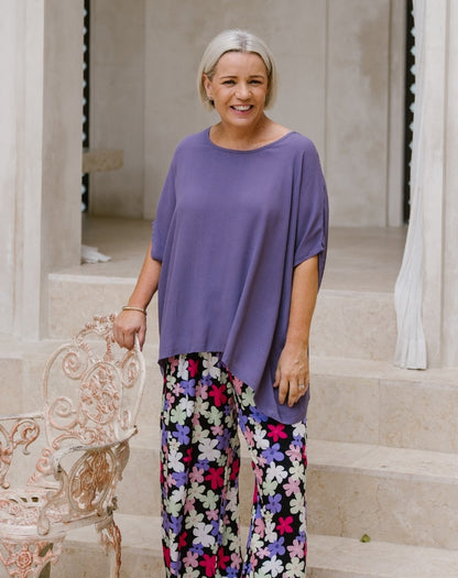 A smiling woman stands on stone steps wearing a loose purple top and colorful floral wide-leg pants, holding a white ornate chair beside her.