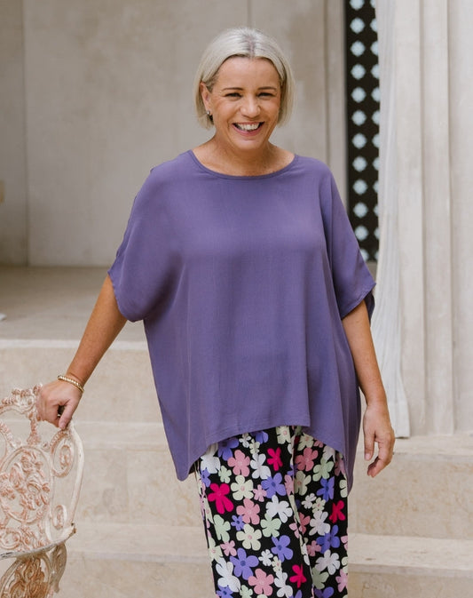 A woman with short gray hair smiling while wearing a loose, short-sleeve purple top and black pants with a colorful floral pattern, standing in a light-colored indoor setting.