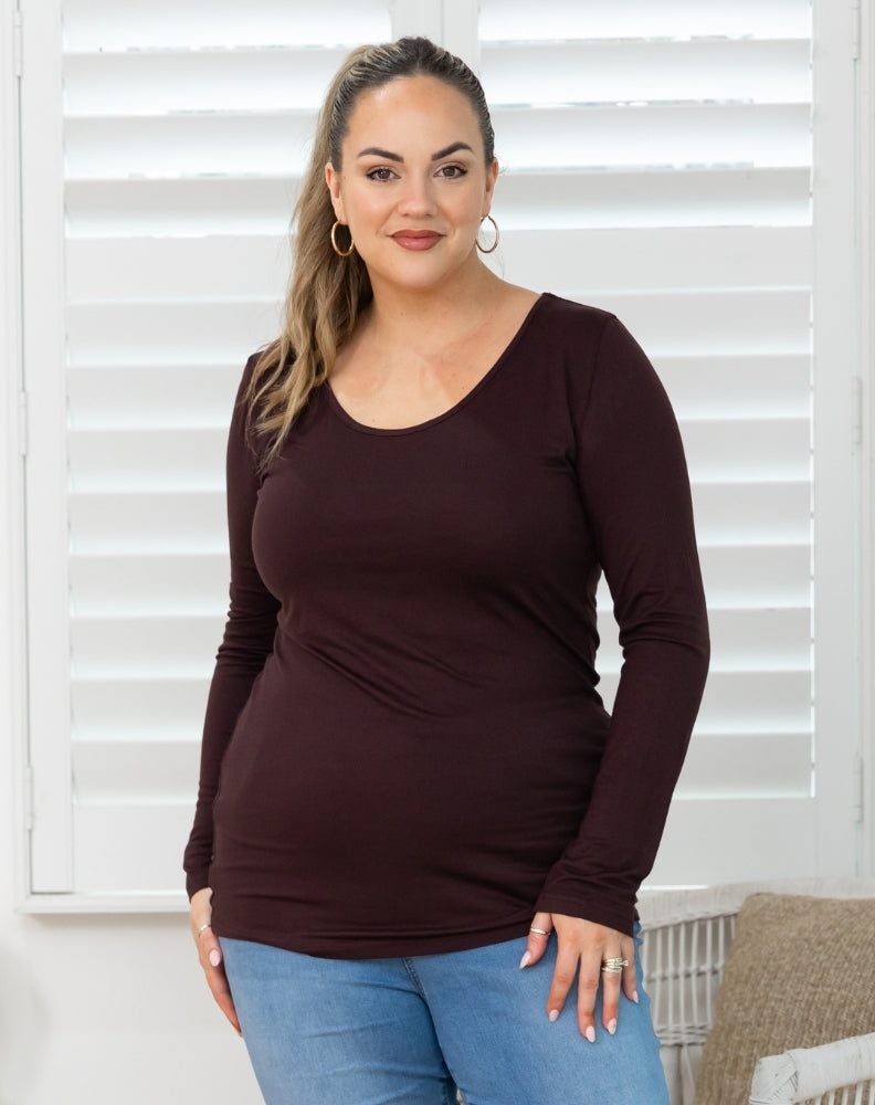 Woman wearing a brown long-sleeve top and blue jeans standing in front of white blinds.