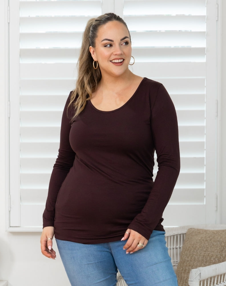 Woman wearing a brown long-sleeve top and blue jeans standing in front of white blinds.