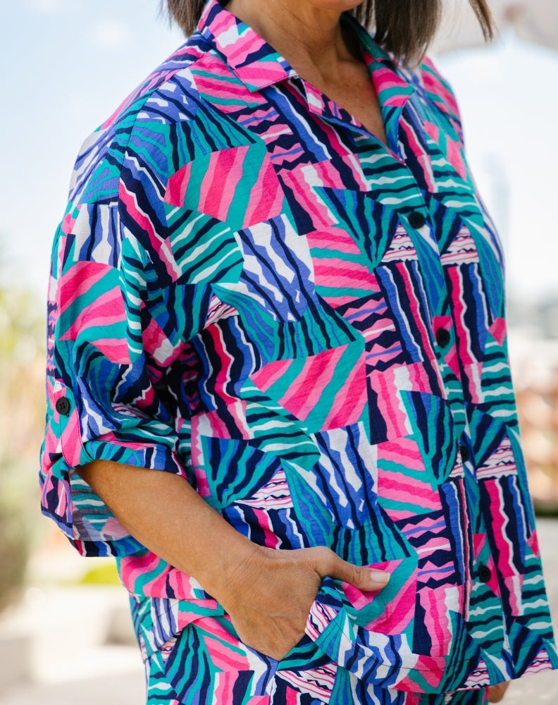 A close-up of a person wearing a short-sleeve button-up shirt with a bold geometric and zebra-like print in bright pink, teal, blue, and white, with one hand in the pocket.