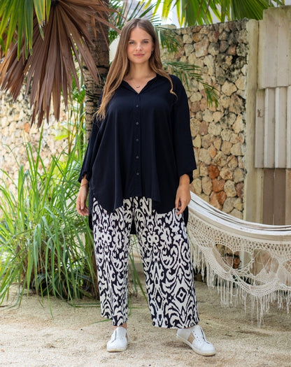 Woman wearing a black top and patterned pants standing in a tropical setting with plants and a hammock.