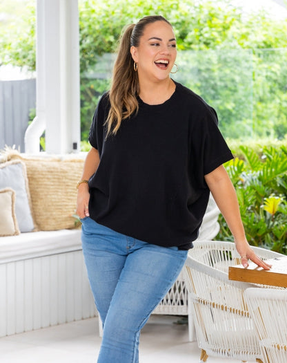 A woman wearing a black short-sleeve t-shirt and light blue jeans stands on a covered patio with green garden foliage in the background.
