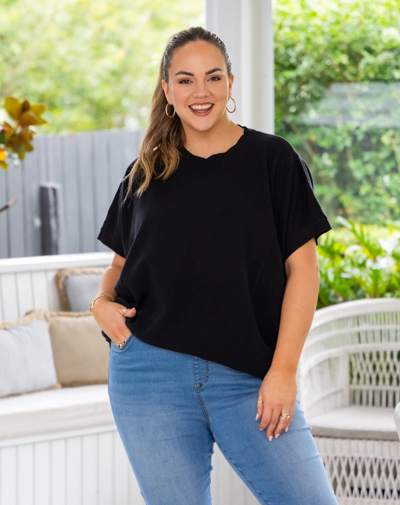 A woman modeling a black short-sleeve crewneck top with scalloped edges, paired with light blue jeans, standing on a white patio with green garden foliage in the background.