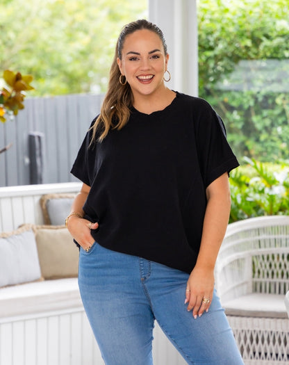 A woman modeling a black short-sleeve crewneck top with scalloped edges, paired with light blue jeans, standing on a white patio with green garden foliage in the background.
