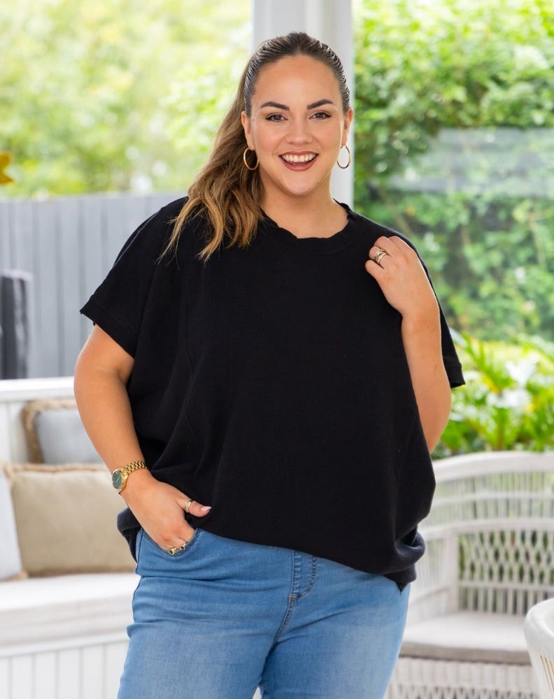 A woman wearing a black short-sleeve crewneck t-shirt and light blue jeans, standing in a bright, modern outdoor patio setting with white wicker furniture and green foliage in the background.