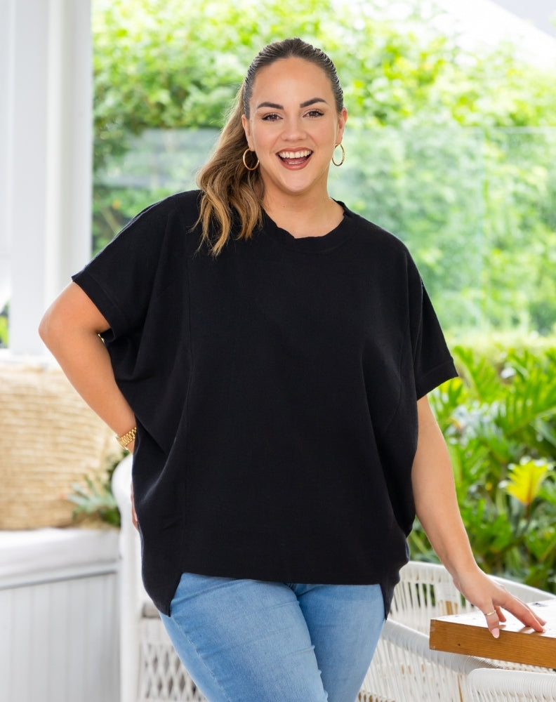 A woman wearing a black short-sleeve tunic top and light blue jeans, standing on a patio with green plants in the background.