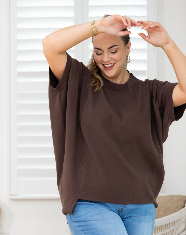 A woman wearing a brown short-sleeve t-shirt and blue jeans, posing in front of white window shutters.