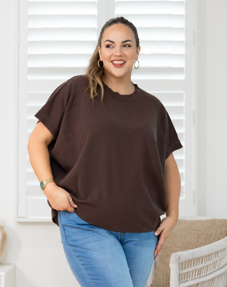 A woman wearing a loose-fitting, short-sleeve brown t-shirt and blue jeans, standing in front of white window shutters.