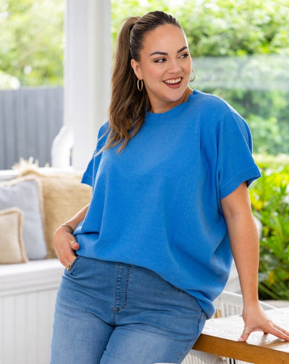 A woman wearing a bright blue oversized short-sleeve t-shirt and blue jeans, leaning on a wooden railing in a bright, sunlit patio setting with green foliage in the background.