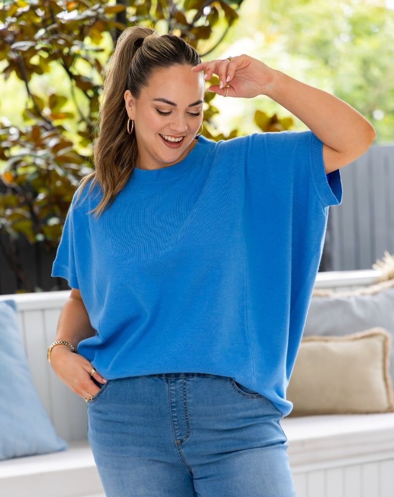 A woman wearing a bright blue short-sleeve knit top and blue jeans, standing outdoors in front of a white bench with cushions and green foliage in the background.