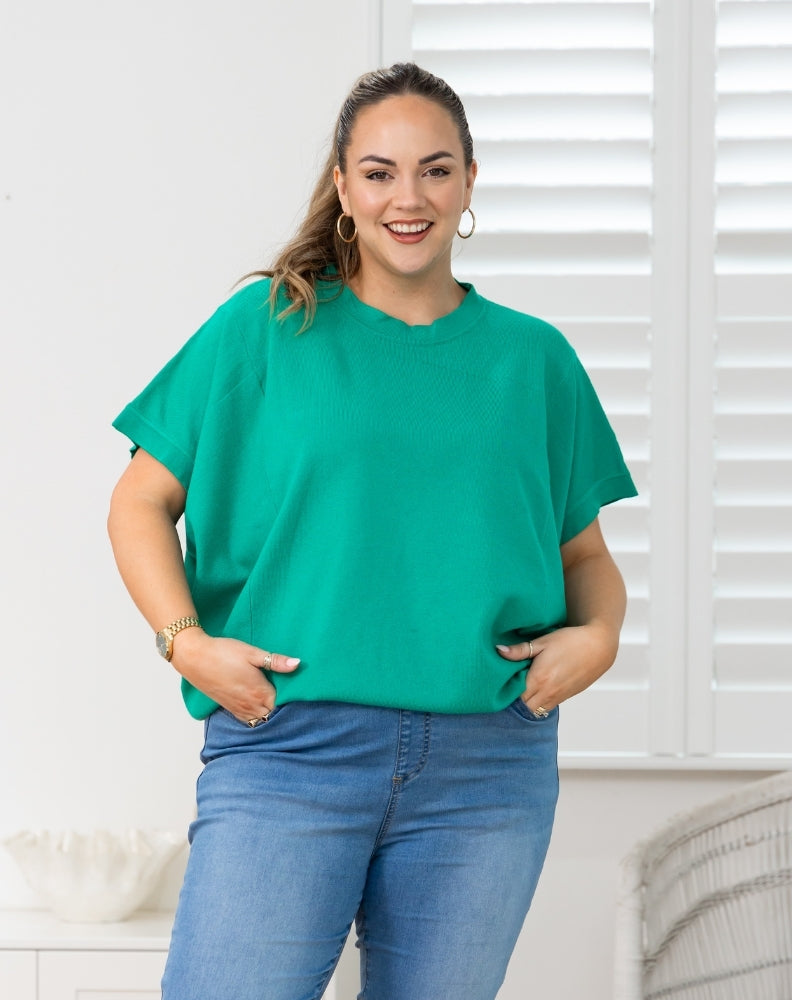 A woman wearing a bright green short-sleeve t-shirt and blue jeans, standing in front of a white shuttered window.