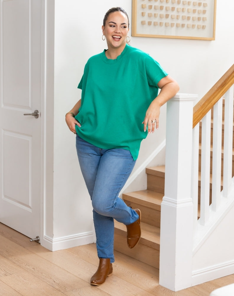 A woman wearing a vibrant green oversized short-sleeve t-shirt, blue jeans, and brown ankle boots is posing on wooden stairs in a bright, modern home interior.