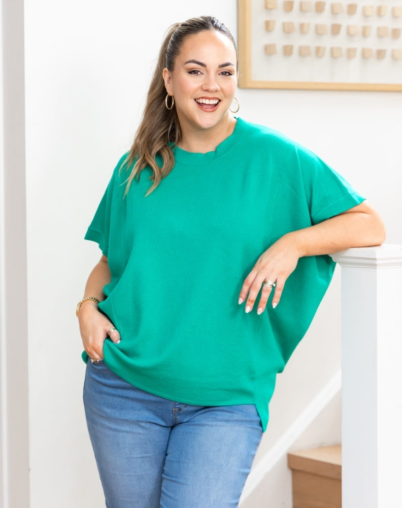 A woman wearing a vibrant green short-sleeve tunic top and blue jeans, posing indoors next to a white staircase railing.