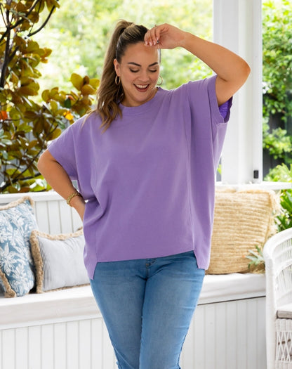 A woman wearing an oversized lavender-purple short-sleeve t-shirt and blue jeans, standing on a white porch with green foliage in the background.