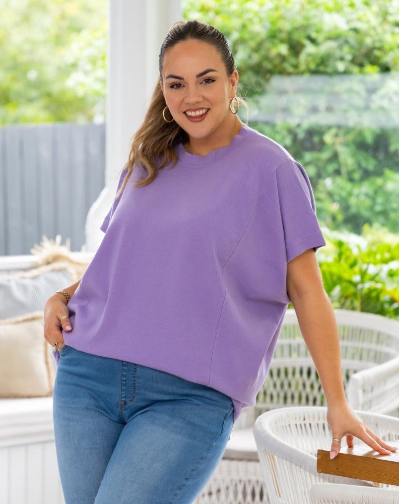 A woman wearing a lavender purple short-sleeve top and blue jeans, standing in a bright, modern outdoor patio setting with white wicker furniture and green foliage in the background.