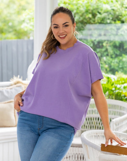 A woman wearing a lavender purple short-sleeve top and blue jeans, standing in a bright, modern outdoor patio setting with white wicker furniture and green foliage in the background.