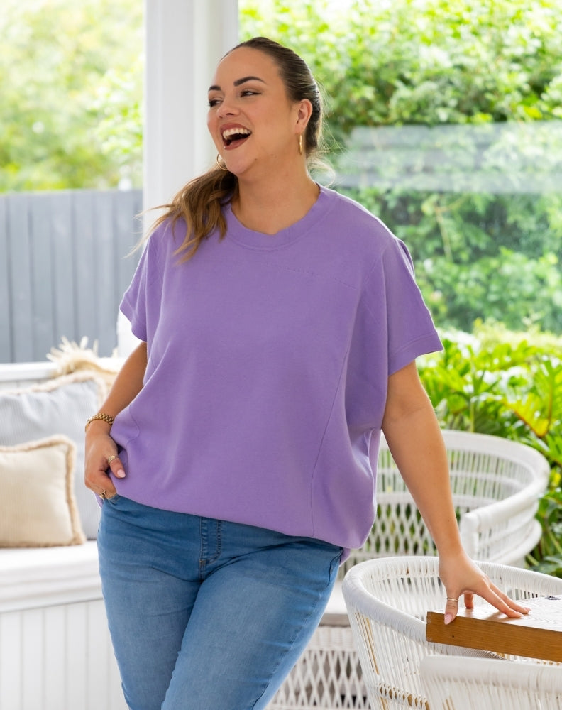 A woman wearing a lavender purple short-sleeve t-shirt and blue jeans, smiling and laughing in an outdoor patio setting with white wicker furniture and green foliage in the background.
