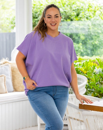 A woman wearing a loose, short-sleeve lavender-purple t-shirt and blue jeans, standing on a white patio with green garden foliage in the background.